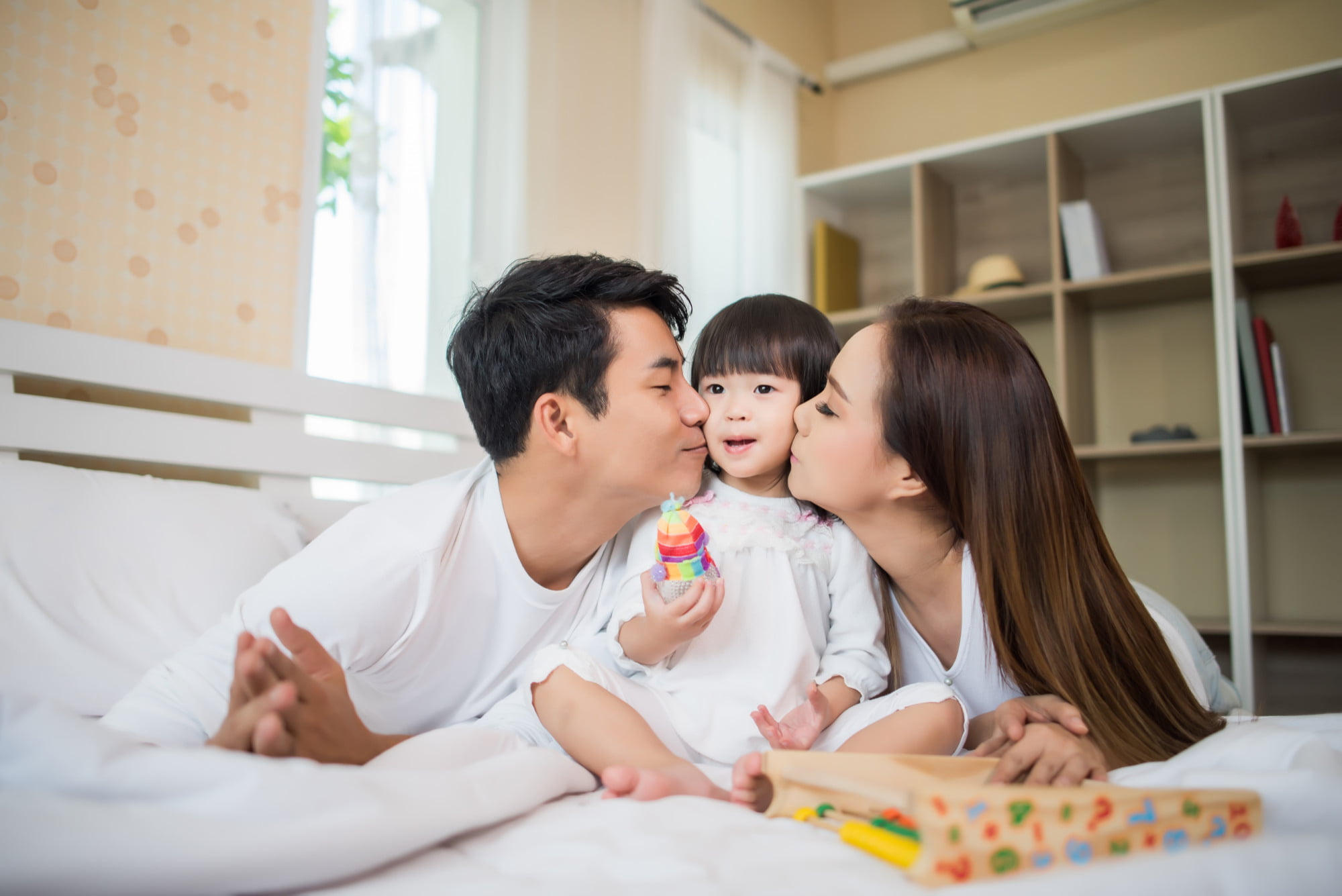 A child sits between two smiling adults on a bed at Star Smiles Pediatric Dentistry in Bloomingdale IL, receiving affectionate kisses on the cheeks. Holding a colorful toy, the child beams with happiness, while a wooden toy tray nearby highlights the warmth and sense of belonging that Apple Health Families: You Belong Here promotes.